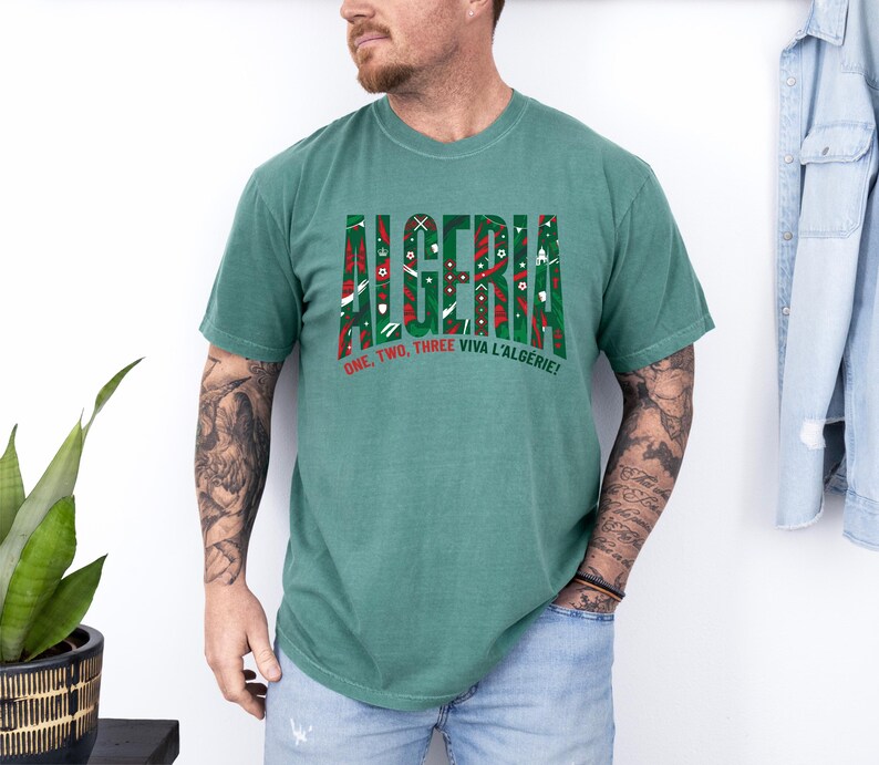 K&ouml;nnte beinhalten: Ein gr&uuml;nes T-Shirt mit dem Wort "ALGERIA" in einer dekorativen Schriftart, gef&uuml;llt mit roten, wei&szlig;en und gr&uuml;nen Mustern. Darunter steht der Satz "ONE, TWO, THREE VIVA L'ALGERIE!" in Rot und Wei&szlig;. Das T-Shirt wird von einer Person getragen.