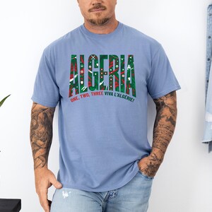 K&ouml;nnte beinhalten: Ein hellblaues T-Shirt mit dem Wort "ALGERIA" in gr&uuml;nen und roten Blockbuchstaben. Das T-Shirt hat auch den Text "ONE, TWO, THREE VIVA L'ALG&Eacute;RIE!" in Rot. Das T-Shirt ist aus weichem Material.