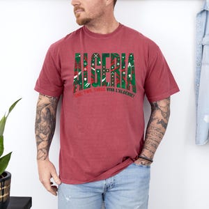 K&ouml;nnte beinhalten: Ein weinrotes T-Shirt mit dem Wort "ALGERIA" in Gr&uuml;n und Rot, mit dekorativen Mustern. Darunter steht der Text "ONE, TWO, THREE VIVA L'ALG&Eacute;RIE!"