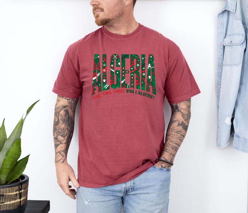 K&ouml;nnte beinhalten: Ein weinrotes T-Shirt mit dem Wort "ALGERIA" in Gr&uuml;n und Rot, mit dekorativen Mustern. Darunter steht der Text "ONE, TWO, THREE VIVA L'ALG&Eacute;RIE!"