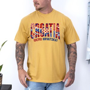 May include: Mustard yellow t-shirt with the word "CROATIA" in red, white, and blue block letters. The text "IDEMO HRVATSKA!" is printed below. The shirt has short sleeves and a crew neck.
