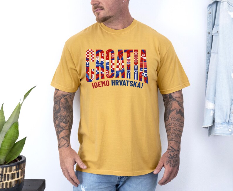 May include: Mustard yellow t-shirt with the word "CROATIA" in red, white, and blue block letters. The text "IDEMO HRVATSKA!" is printed below. The shirt has short sleeves and a crew neck.