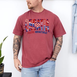 May include: A maroon t-shirt with the word "CROATIA" in a multi-colored design featuring Croatian flags and symbols. Below the word is the phrase "IDEMO HRVATSKA!" in blue. The shirt is short-sleeved.