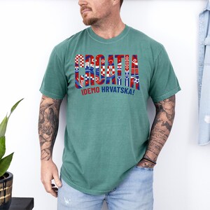 May include: A seafoam green t-shirt with the word "CROATIA" in a colorful, flag-themed design. Below the word is the phrase "IDEMO HRVATSKA!" in red and white. The shirt is worn by a person.