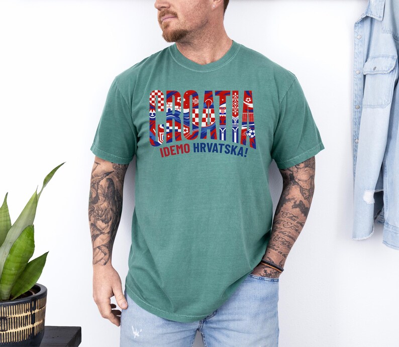 May include: A seafoam green t-shirt with the word "CROATIA" in a colorful, flag-themed design. Below the word is the phrase "IDEMO HRVATSKA!" in red and white. The shirt is worn by a person.