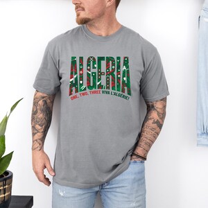 K&ouml;nnte beinhalten: Ein graues T-Shirt mit dem Wort "ALGERIA" in gr&uuml;nen und roten Blockbuchstaben. Das Shirt hat auch den Text "ONE, TWO, THREE VIVA L'ALG&Eacute;RIE!" in Rot und Wei&szlig;. Das T-Shirt wird von einer Person getragen.