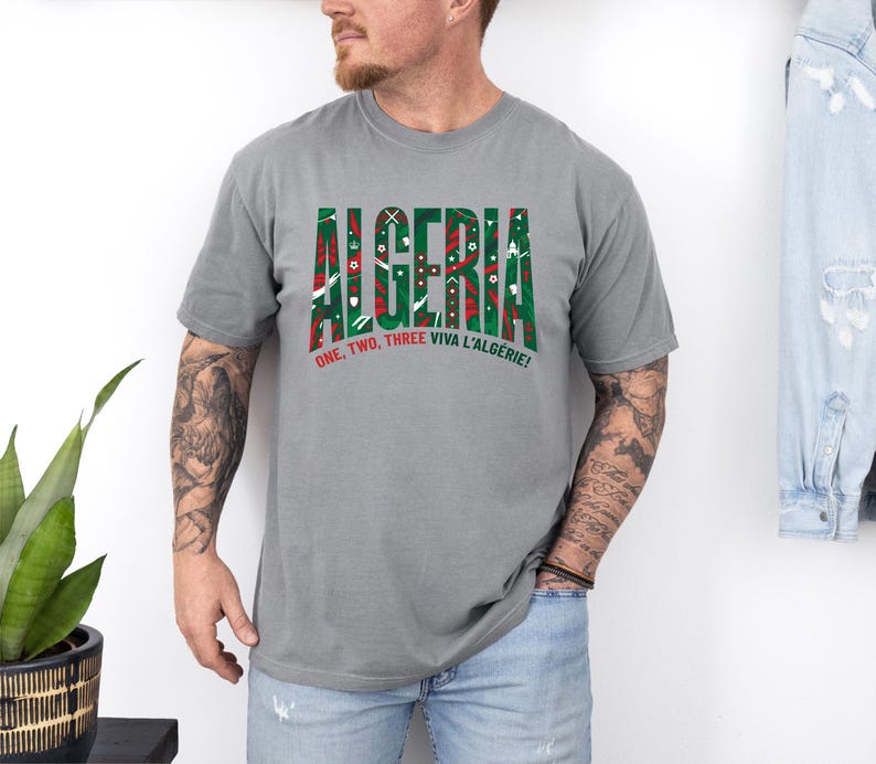 K&ouml;nnte beinhalten: Ein graues T-Shirt mit dem Wort "ALGERIA" in gr&uuml;nen und roten Blockbuchstaben. Das Shirt hat auch den Text "ONE, TWO, THREE VIVA L'ALG&Eacute;RIE!" in Rot und Wei&szlig;. Das T-Shirt wird von einer Person getragen.