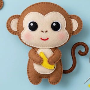 May include: A handmade felt monkey toy in brown, cream, and yellow. The monkey has large black eyes, rosy cheeks, and a red smile. It is holding a yellow banana. The background is a light blue.