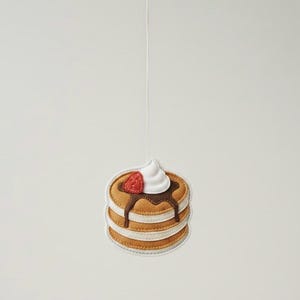 Può includere: Un ornamento di feltro a forma di pancake con tre pancake impilati, sormontati da panna montata, una fetta di fragola e sciroppo al cioccolato. L'ornamento è sospeso da un sottile filo bianco, perfetto per le decorazioni.