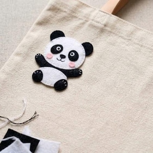 Op de afbeelding: Een handgemaakte panda beer vilt applicatie in zwart, wit en roze is bevestigd aan een beige canvas tote bag. De panda heeft zwarte oren, ogen en poten, met roze wangen. De tas heeft bruine leren handvatten.