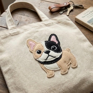 Op de afbeelding: Een beige canvas draagtas met een viltapplicatie van een Franse Bulldog. De hond heeft een zwart-witte kop, een bruin lichaam en roze oren. De tas heeft een eenvoudig ontwerp met twee handvatten.