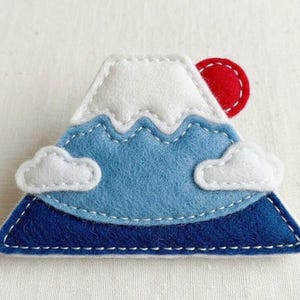 Peut inclure: Applique en feutre en forme de montagne, avec un sommet enneigé blanc, des pentes bleues et une base bleu foncé. Deux nuages blancs et un soleil rouge sont également inclus. L'applique est cousue à la main avec du fil blanc.