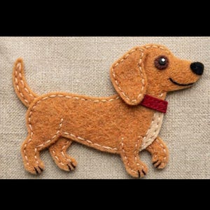 Pode incluir: Aplique de feltro de um cão dachshund em tons de castanho, com coleira vermelha e nariz preto. O cão é costurado com linha branca, e o fundo é um tecido neutro.
