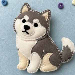 Puede incluir: Un perro de fieltro hecho a mano en posición sentada, con una combinación de colores gris y blanco, ojos y nariz negros. El perro tiene una cola esponjosa y está cosido con hilo blanco. El fondo es azul claro con pequeños círculos de fieltro.