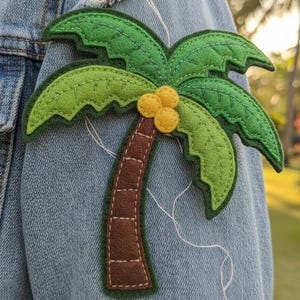 Könnte beinhalten: Ein Filz-Palmen-Aufnäher in Grün-, Braun- und Gelbtönen, auf eine Jeansjacke genäht. Der Aufnäher hat ein detailliertes Design mit einem braunen Stamm, grünen Wedeln und gelben Kokosnüssen. Der Aufnäher ist etwa 13 cm breit.