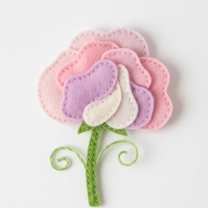 Könnte beinhalten: Eine handgefertigte Filzblume in Rosa-, Lavendel- und Cremetönen mit einem grünen Stiel und zarten Stickdetails. Die Blütenblätter sind geschichtet und genäht, wodurch ein dreidimensionaler Effekt entsteht. Der Stiel hat gestickte grüne Wirbel.