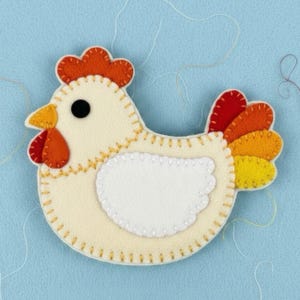 Puede incluir: Un pollo de fieltro hecho a mano con cuerpo color crema, vientre blanco y detalles en naranja, rojo y amarillo. El pollo tiene un ojo negro y está cosido con hilo amarillo. El fondo es azul claro.