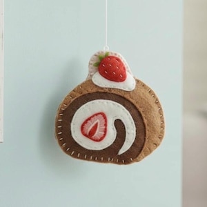 Puede incluir: Adorno de fieltro con forma de brazo de gitano. El bizcocho marrón tiene un relleno de crema blanca con una rodaja de fresa roja en el centro. Una fresa entera con hojas verdes está encima. El adorno está suspendido por un hilo blanco.