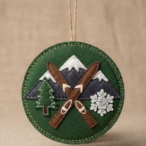 May include: A round, green felt ornament with a winter theme. It features a mountain scene with white snow-capped peaks, a green tree, and brown skis crossed over each other. A white snowflake is also included.