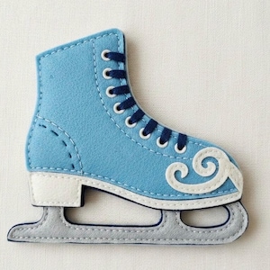 May include: A light blue felt ice skate with white detailing and dark blue laces. The skate has a decorative white swirl and a grey blade. The stitching is visible, giving a handcrafted appearance.