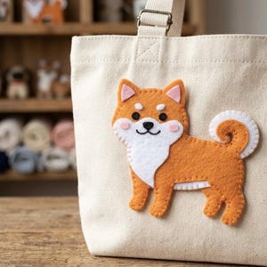 Puede incluir: Bolso tote de lona beige con una aplicación de fieltro de un perro Shiba Inu. El perro es naranja, blanco y rosa, con una cola rizada. El bolso tiene una correa de color crema.