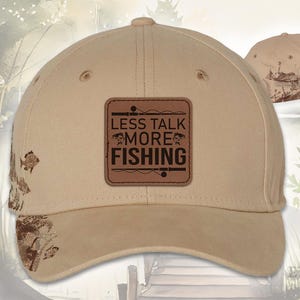 May include: Tan baseball cap with a brown leather patch that reads "LESS TALK MORE FISHING". The cap has a curved brim and a fishing-themed design on the side. The cap is made of a durable fabric.