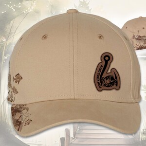 May include: Tan baseball cap with a brown leather patch featuring a fish hook and a fish graphic, with the text "FISH VIBES ONLY." The cap has a curved brim and a decorative fish and aquatic plant design on the side.
