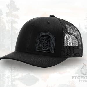 May include: Black trucker hat with a mesh back and a curved brim. The front features a rectangular patch with a Grim Reaper design and the text "MAY THE BRIDGES I BURN LIGHT THE WAY."