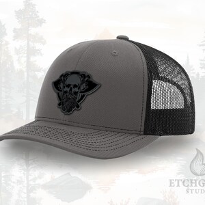 May include: Gray trucker hat with a black mesh back and a skull patch. The patch features a skull with a beard and wings. The hat has a curved brim and a snapback closure. The hat is a stylish accessory.