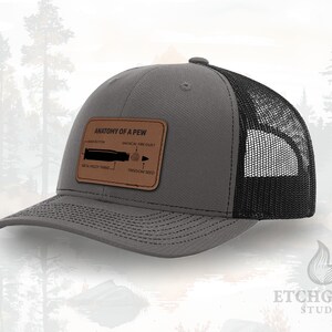 May include: Gray trucker hat with a black mesh back and a curved brim. The hat features a brown leather patch with the text "ANATOMY OF A PEW" and an illustration of a bullet with labeled parts. The hat is a casual accessory.