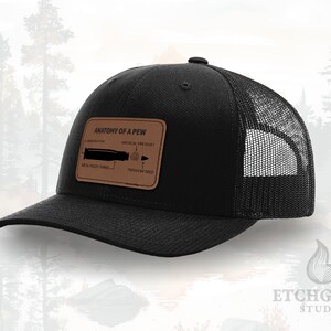 May include: Black trucker hat with a mesh back and a curved brim. The hat features a brown leather patch with the text "ANATOMY OF A PEW" and an illustration of a bullet. The hat is a classic style, suitable for outdoor activities.