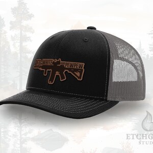 May include: Black and gray trucker hat with a brown leather patch featuring a rifle graphic and the text "RED WHITE AND PEWPEW". The hat has a curved brim and a mesh back.
