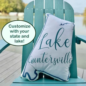 May include: A white pillow with blue text that reads "Lake Guntersville" on a teal adirondack chair. The pillow is on a wooden deck with a lake in the background.