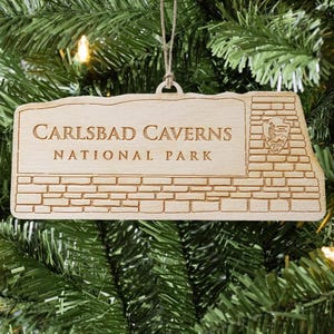 May include: Wooden Carlsbad Caverns National Park ornament. The rectangular ornament features the park's name and logo, with a brick-like design. It is suspended by a natural twine string, perfect for holiday decorating. The ornament is made of light-colored wood.