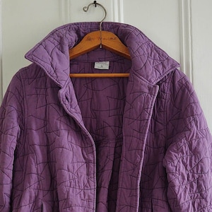 May include: A quilted purple jacket with a geometric pattern, hanging on a wooden hanger. The jacket has a wide collar, two pockets, and a button closure. The jacket is approximately 28 inches long.