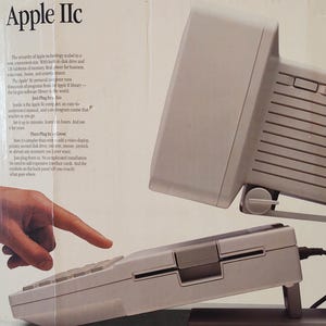 以下が含まれることがあります： Apple IIcパーソナルコンピュータの段ボール箱。Appleのロゴと製品名が記載されています。箱にはベージュのコンピュータキーボードユニット、モニター、プリンター、ディスクドライブ、モデムなどの周辺機器のアイコンが表示されています。