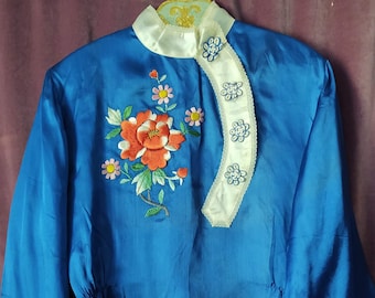 May include: A vibrant blue satin blouse with long sleeves and a mandarin collar. The blouse features an embroidered floral design in shades of red, orange, pink, and green. The collar and trim are white with delicate detailing.