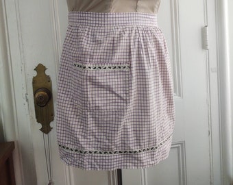 Op de afbeelding: Een vintage schort op een mannequin. Het schort heeft een paars en wit geruit patroon, een zak en decoratieve rand. De buste van de mannequin is lichtbruin.