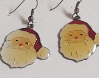 Puede incluir: Un par de pendientes colgantes con un diseño de cara de Papá Noel con gorro rojo, barba color crema y ganchos de metal plateado. Las caras de Papá Noel están hechas en esmalte con contornos dorados, mostrando un motivo navideño clásico.