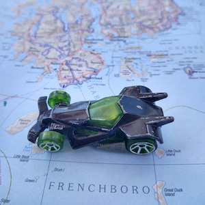 May include: A dark gray and green toy car with green wheels sits on a map. The car has a futuristic design with a transparent green cockpit. The map shows a coastal area with islands and the text "FRENCHBORO".