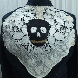 May include: Dark denim jacket with a large white lace appliqué on the back. A black skull is centered on the lace, with a gold trim detail. The lace design extends across the shoulders.