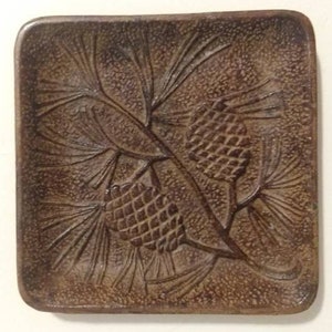 May include: A square, brown ceramic tray with a raised design of pine branches and pine cones.