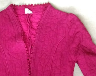 Puede incluir: Chaqueta acolchada magenta con mangas largas. La chaqueta tiene cuello en V y un ribete festoneado a lo largo de los bordes. La tela tiene un patrón acolchado texturizado. La chaqueta está abierta por delante.