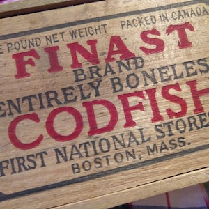 Könnte beinhalten: Eine Holzkiste mit schwarzer und roter Beschriftung. Die Kiste trägt die Aufschrift "One pound net weight packed in Canada, Finast Brand, Entirely Boneless Codfish, First National Stores Inc, Boston, Mass."