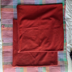 May include: Two square, solid red pillow covers are stacked on top of each other. The pillow covers are made of a soft fabric. The pillows are placed on a colorful plaid fabric.