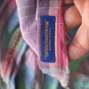 K&ouml;nnte beinhalten: Nahaufnahme eines karierten Hemdes in Rosa-, Lila- und Gr&uuml;nt&ouml;nen. Das Hemd hat ein blaues Etikett mit der Aufschrift "HIGH GRADE WESTERN WEAR" in goldfarbenen Buchstaben. Der Stoff ist weich und scheint Baumwolle zu sein.
