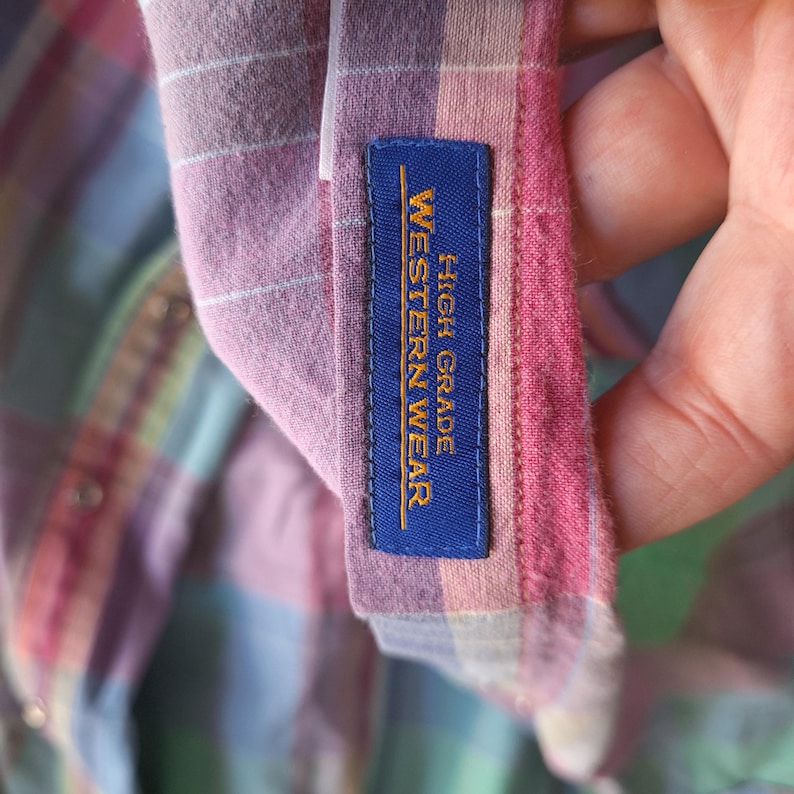 K&ouml;nnte beinhalten: Nahaufnahme eines karierten Hemdes in Rosa-, Lila- und Gr&uuml;nt&ouml;nen. Das Hemd hat ein blaues Etikett mit der Aufschrift "HIGH GRADE WESTERN WEAR" in goldfarbenen Buchstaben. Der Stoff ist weich und scheint Baumwolle zu sein.