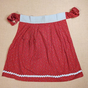 Puede incluir: Un delantal rojo con un estampado floral y una cinturilla azul claro. El delantal tiene un ribete blanco festoneado a lo largo del borde inferior y lazos de tela a juego. La tela es de algodón ligero.