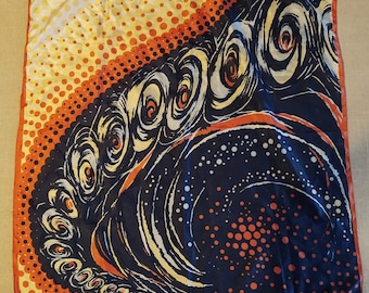 May include: Square silk scarf with a vibrant abstract design. The scarf features a gradient of yellow and orange dots, transitioning to a navy blue background with swirling white and orange patterns. The edges are trimmed with a red border.