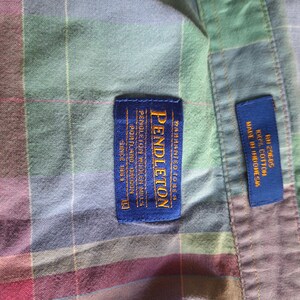 K&ouml;nnte beinhalten: Nahaufnahme eines karierten Hemdes mit einem blauen Pendleton-Etikett. Das Hemd ist eine Mischung aus Farben, darunter Gr&uuml;n, Rot und Blau. Auf dem Etikett steht "Pendleton Woolen Mills, Portland Oregon, Since 1863". Ein kleineres blaues Etikett lautet "Made in Indonesia, 100% Cotton".